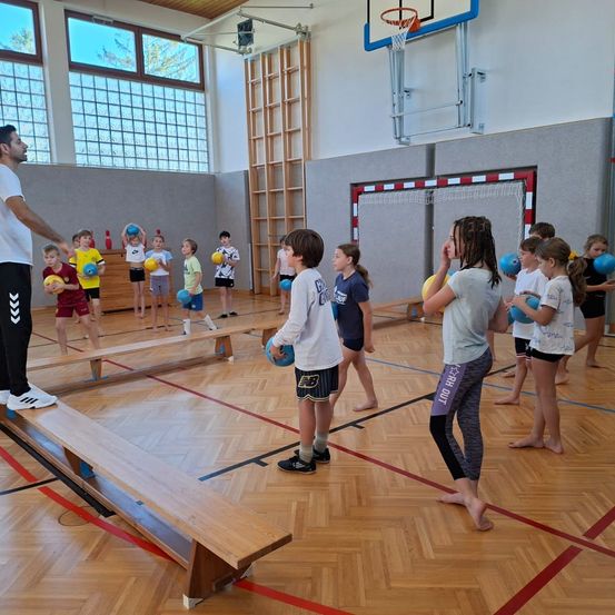 In einer Turnhalle spielen eine Gruppe von Kindern und ein Erwachsener mit Bällen. Sie stehen auf Bänken und halten Bälle, wahrscheinlich spielen sie ein Spiel. Die Turnhalle hat Holzböden, einen Basketballkorb und Glasfenster.