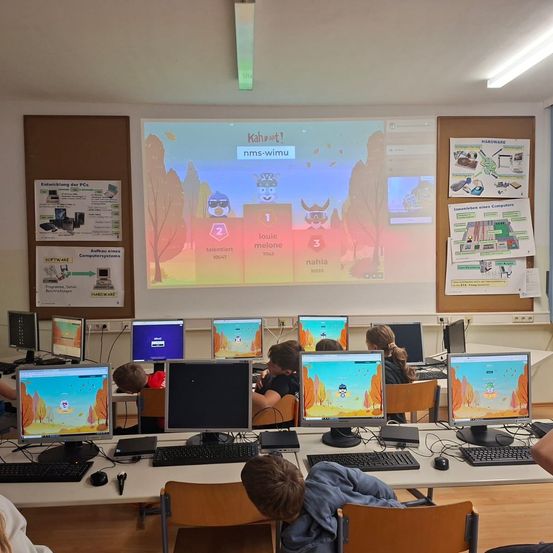 Ein Klassenzimmer mit Schülern vor Computern, eine Projektionsfläche zeigt ein Spiel mit Charakteren und Punkten. Poster an den Wänden liefern Informationen.
