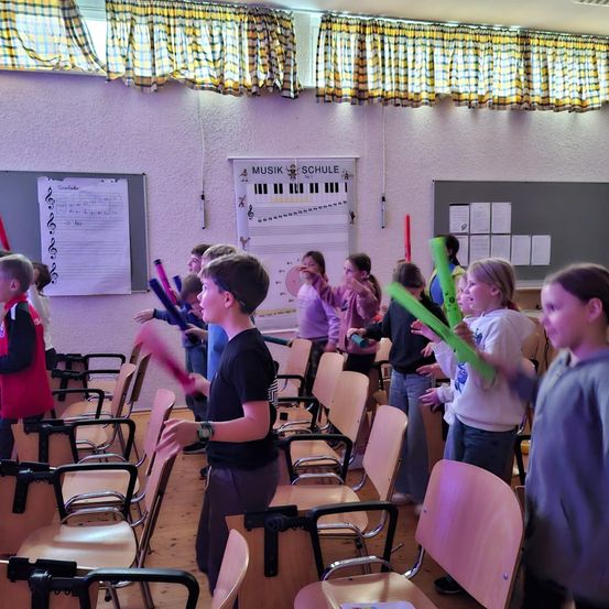 Eine Gruppe von Kindern in einem Klassenzimmer hält und spielt bunte Musikinstrumente. Ein Musiknotenposter ist an der Wand angebracht.