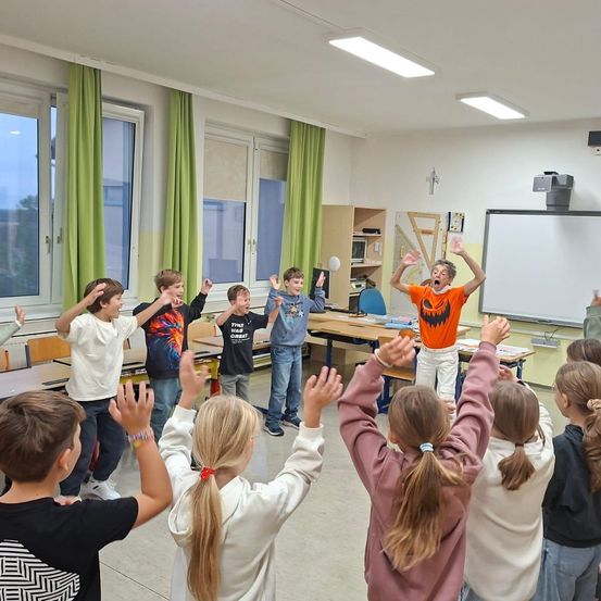 Ein Klassenzimmer voller Kinder, die mit erhobenen Händen tanzen. Ein Junge in einem orangefarbenen Shirt lächelt und springt in der Mitte. Der Raum hat weiße Wände, grüne Vorhänge und eine Tafel.