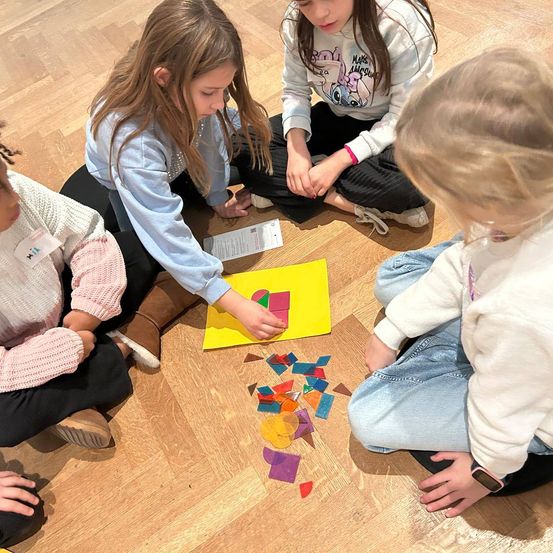 Vier junge Mädchen sitzen auf einem Holzboden, möglicherweise in einem Klassenzimmer. Sie sind in einer Aktivität mit farbenfrohen geometrischen Formen beschäftigt. Ein Mädchen zeigt auf ein gelbes Stück.