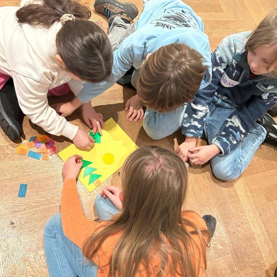 Vier Kinder arbeiten gemeinsam an einem Bastelprojekt und ordnen bunte Formen auf einer gelben Papierbasis an. Jedes Kind konzentriert sich auf seine Aufgabe, wobei eines ein grünes Stück verwendet.