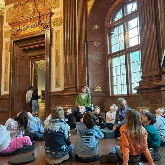 Eine Gruppe von Kindern sitzt auf dem Boden in einem großen Raum und hört einer Frau zu. Eine offene Tür zeigt einen weiteren Raum.