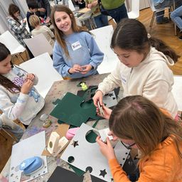 Mehrere junge Mädchen arbeiten an einem Bastelprojekt. Sie sitzen um einen Tisch herum mit grünen Papierausschnitten und verschiedenen Bastelutensilien. Eines der Mädchen lächelt und hält eine Schere. Die anderen konzentrieren sich auf ihre Aufgaben.