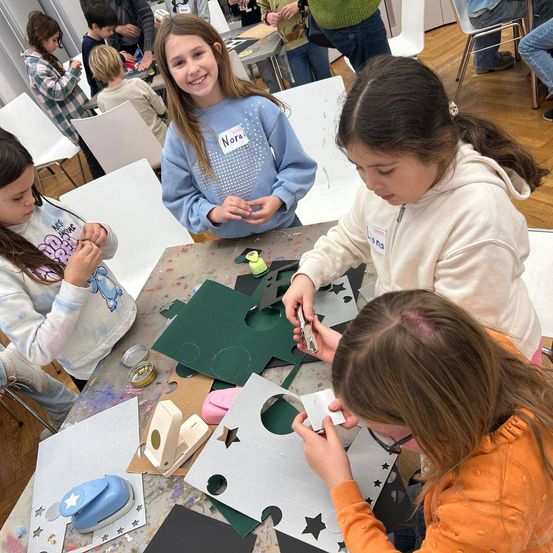 Mehrere junge Mädchen arbeiten an einem Bastelprojekt. Sie sitzen um einen Tisch herum mit grünen Papierausschnitten und verschiedenen Bastelutensilien. Eines der Mädchen lächelt und hält eine Schere. Die anderen konzentrieren sich auf ihre Aufgaben.