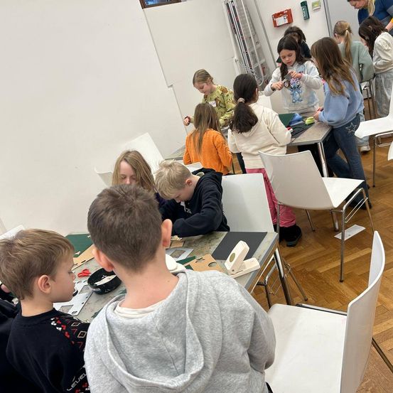 Eine Gruppe von Kindern in einem Workshop, einige sitzen an Tischen, andere stehen. Einige arbeiten an Papierbasteleien, während andere sich unterhalten. Der Raum hat weiße Wände und Holzböden.