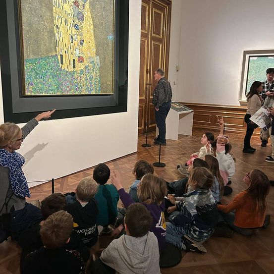 Eine Gruppe von Kindern sitzt auf dem Boden und beobachtet eine Frau, die auf ein Gemälde an der Wand zeigt.