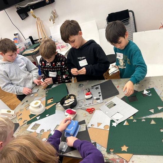 Fünf Kinder sind um einen Tisch mit Papierbasteleien versammelt. Sie scheinen an einem Projekt zu arbeiten, mit verschiedenen Materialien wie Scheren, Papier und Aufklebern. Die Kinder tragen Freizeitkleidung. Im Hintergrund befindet sich eine Wand mit einigen Dekorationen.