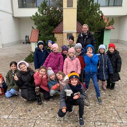 Eine Gruppe von Kindern und Erwachsenen posiert für ein Foto in einem verschneiten Außenbereich, mit zwei kleinen Gebäuden im Hintergrund.