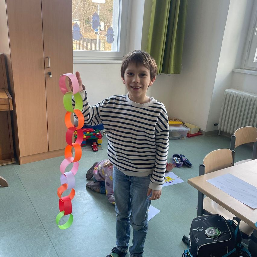 Ein junger Junge in einem gestreiften Shirt hält eine bunte Papierkette in einem Klassenzimmer mit Fenster und Heizkörper hoch.