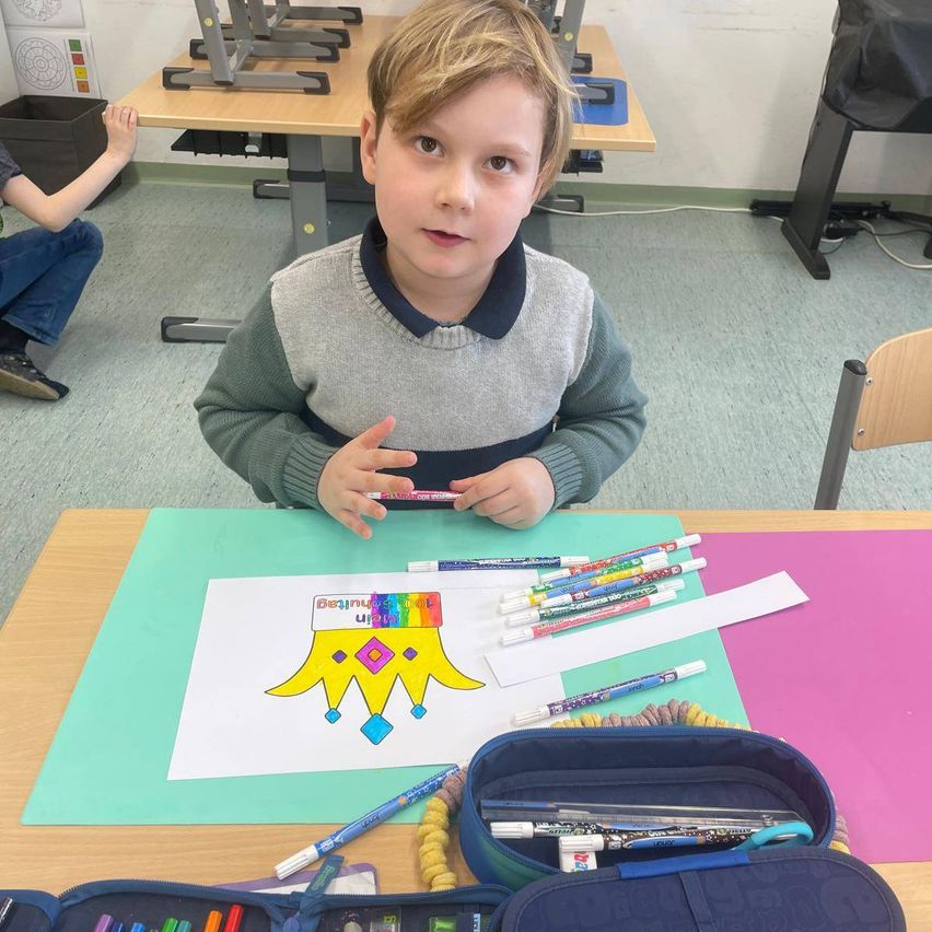 Ein Junge sitzt in einem Klassenzimmer an einem Schreibtisch und hält einen Stift, während er eine Zeichnung ausmalt. Er ist umgeben von einem Bleistiftetui, einer Tasche und verschiedenen farbigen Stiften.