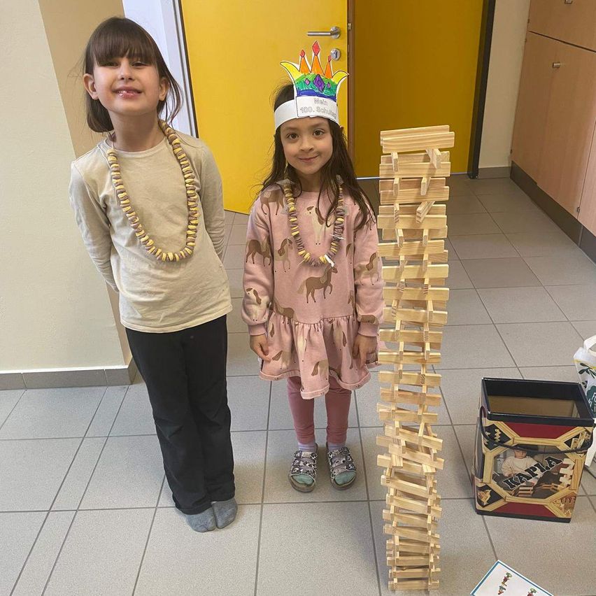 Zwei junge Mädchen stehen neben einem hohen Turm aus Holzklötzen. Ein Mädchen trägt eine Kronen-Kopfbedeckung und eine Halskette. Der Raum hat gelbe Wände und einen Fliesenboden.