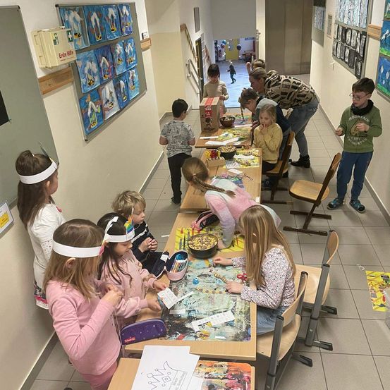 Kinder, die Stirnbänder tragen, versammeln sich um einen Tisch in einem Klassenzimmer. Ein Kind greift nach einer Schüssel mit Snacks. Auf dem Tisch befinden sich mehrere Kunstmaterialien.
