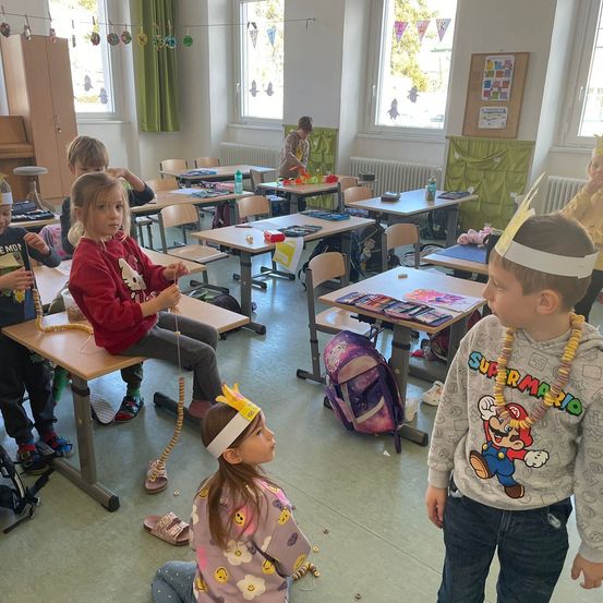 Ein Klassenzimmer mit Kindern, die Stirnbänder tragen. Ein Junge steht und schaut auf ein Mädchen, das sitzt und eine Schnur mit Perlen hält. Der Raum hat Schreibtische, Stühle und einen Schrank. Es gibt Fenster mit Vorhängen und eine Pinnwand.