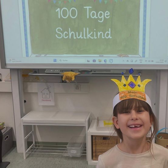 Ein junges Mädchen im Klassenzimmer mit einer Krone, auf der 100 Tage Schulkind steht. Dahinter ist ein Projektionsbildschirm mit denselben Worten.