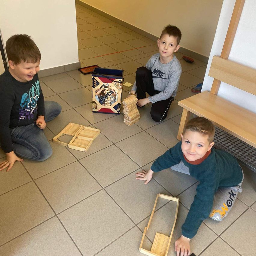 Drei Jungen spielen mit Holzklötzen auf einem gefliesten Boden, ein Junge kniet und lächelt, ein anderer krabbelt auf dem Boden, und ein dritter sitzt und lächelt. Eine Holzbank und eine Aufbewahrungsbox sind in der Nähe.