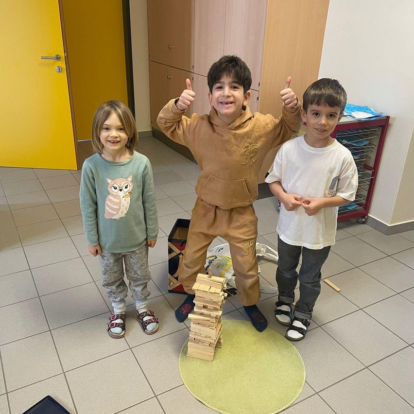 Drei Kinder in einem Raum, eines hält einen Stapel Blöcke, gibt Daumen hoch, trägt Freizeitkleidung und steht auf gefliesten Böden.