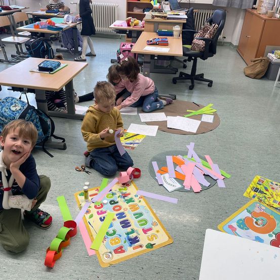 Kinder in einem Klassenzimmer sind an einer Kunstaktivität mit Papieren und Scheren beteiligt, ein Junge lächelt und hält eine Kette aus Papier.