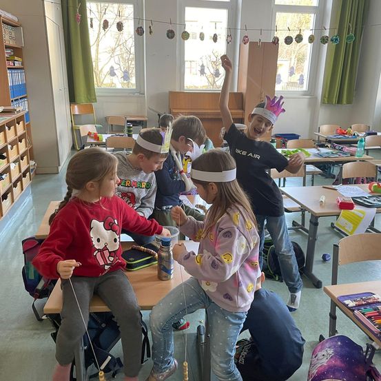 Eine Gruppe von Kindern in einem Klassenzimmer mit Kopfbändern spielt mit einem Ball an einer Schnur. Sie sitzen an Tischen mit verschiedenen Gegenständen darauf.