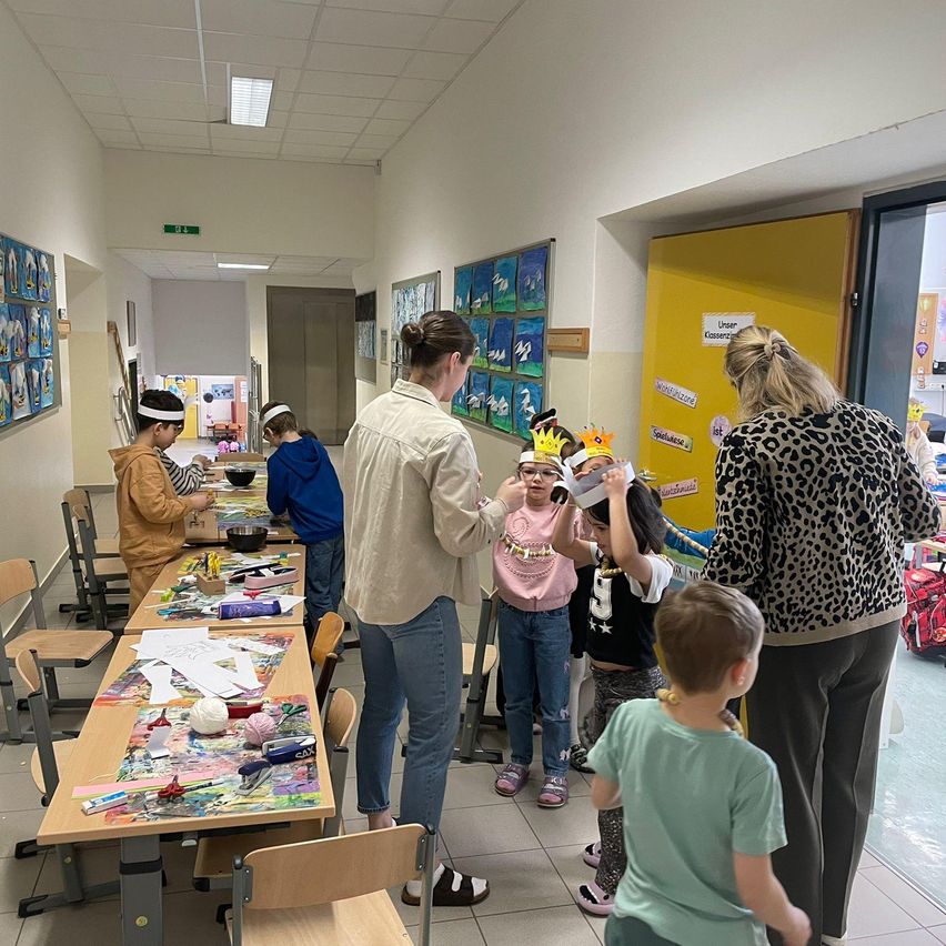 Eine Gruppe von Kindern mit Kronen steht in einem Flur, zwei Frauen sind in der Nähe. Einige sitzen auf Stühlen, und ein Tisch hat verschiedene Bastelmaterialien.