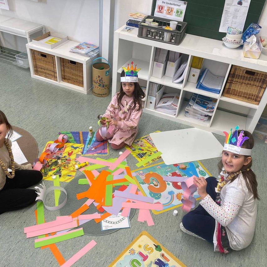 Drei junge Mädchen in einem Klassenzimmer sitzen auf dem Boden, tragen Papierkronen und beschäftigen sich mit einer Bastelaktivität. Sie sind von verschiedenen farbenfrohen Papieren und Materialien umgeben. Dahinter steht ein weißer Regal, das Bücher und Aufbewahrungsboxen enthält.