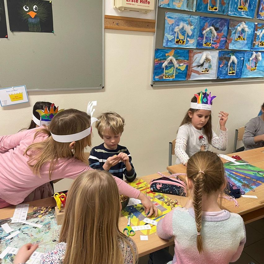 Mehrere Kinder in einem Klassenzimmer tragen Stirnbänder und arbeiten an einem Bastelprojekt. Eine Tafel und Kunstwerke sind an der Wand angebracht.