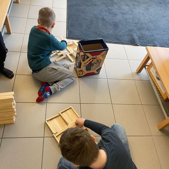 Zwei junge Jungen spielen mit Holzklötzen auf einem gefliesten Boden. Ein Junge trägt einen grünen Pullover und der andere ein schwarzes T-Shirt. Ein Aufbewahrungsbehälter und ein Stapel von Blöcken sind in der Nähe.