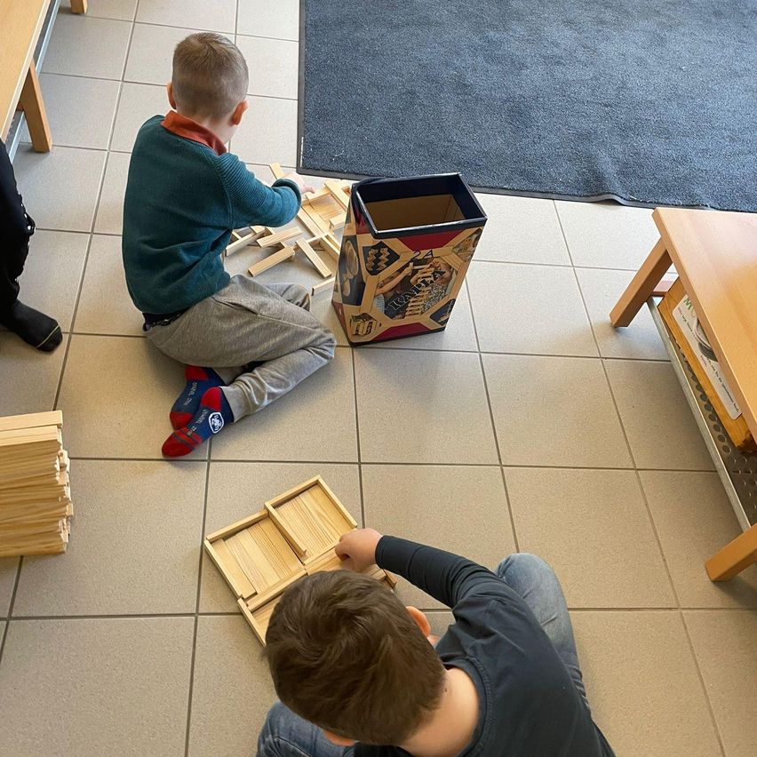Zwei junge Jungen spielen mit Holzklötzen auf einem gefliesten Boden. Ein Junge trägt einen grünen Pullover und der andere ein schwarzes T-Shirt. Ein Aufbewahrungsbehälter und ein Stapel von Blöcken sind in der Nähe.