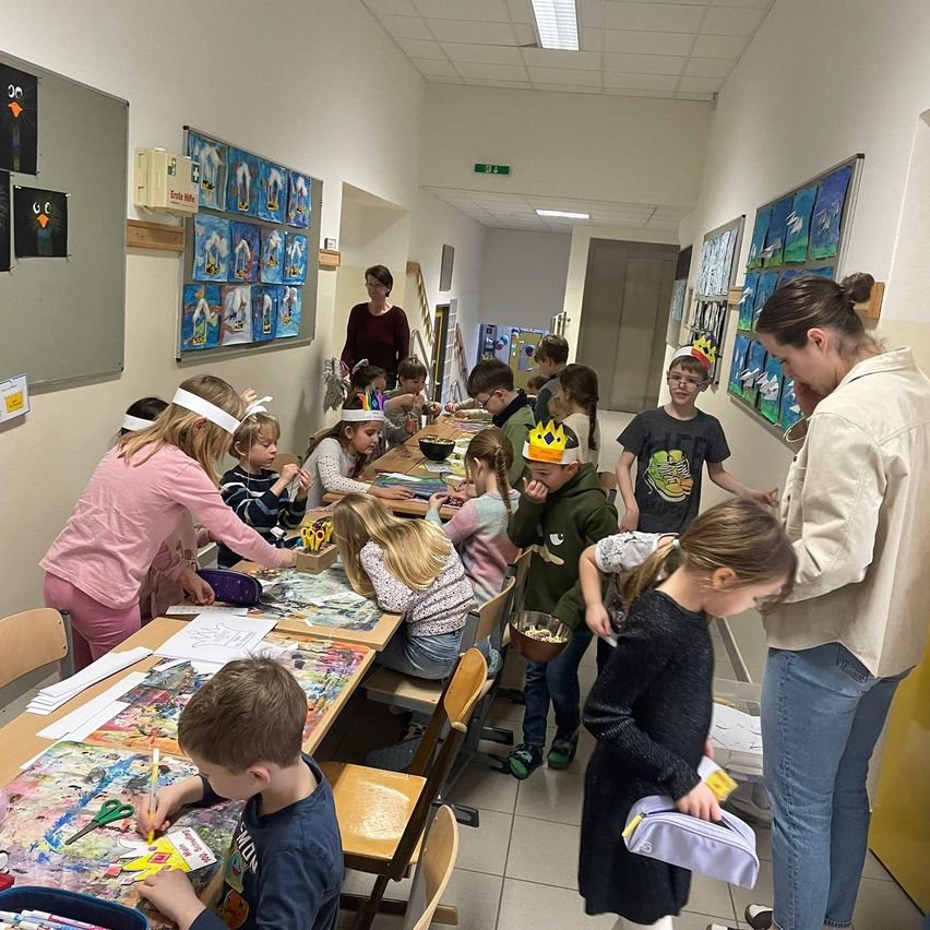 Eine Gruppe von Kindern und Erwachsenen befindet sich in einem Raum, in dem sie Kunst und Handwerk betreiben. Einige Kinder tragen Stirnbänder und Masken. Eine Frau hält einen Stift. Der Raum hat Gemälde an den Wänden und eine Tür am Ende.