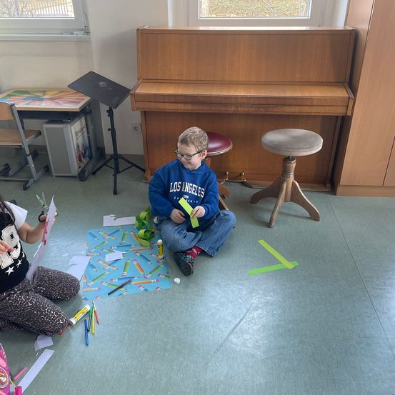 Zwei Kinder sitzen auf dem Boden und machen eine Bastelaktivität. Sie sind von bunten Papieren, Stiften und Aufklebern umgeben. Dahinter steht ein Holzklavier und ein Hocker.