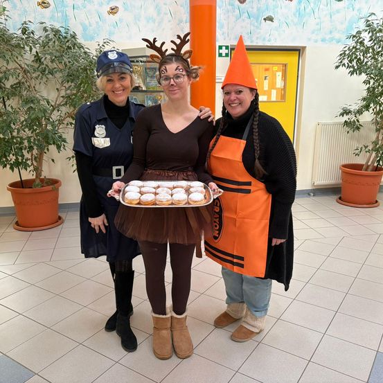 Drei Frauen in Kostümen, eine hält eine Tablett mit Donuts. Eine Frau ist als Polizistin, eine als Rentier und eine als Buntstift verkleidet.