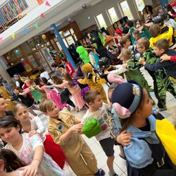 Eine große Gruppe von Kindern in verschiedenen Kostümen tanzt in einem Schulflur. Einige tragen Tierkostüme, während andere Superhelden-Outfits tragen. Der Raum ist mit bunten Wimpelketten dekoriert.