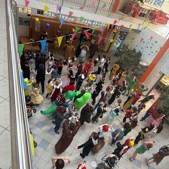 Eine große Gruppe von Kindern in Kostümen tanzt in einem Schulflur mit bunten Dekorationen.