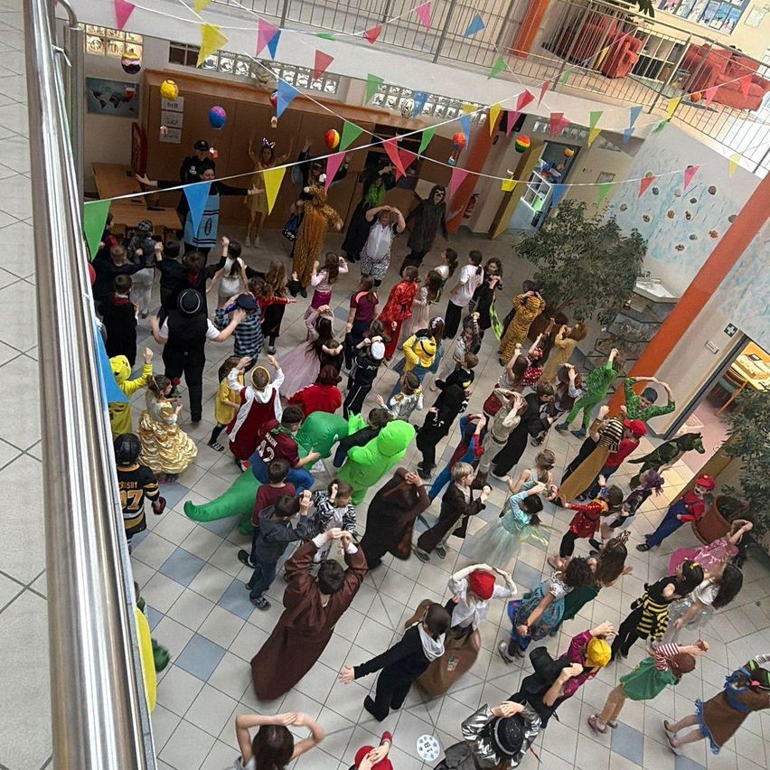 Eine große Gruppe von Kindern in Kostümen tanzt in einem Schulflur mit bunten Dekorationen.