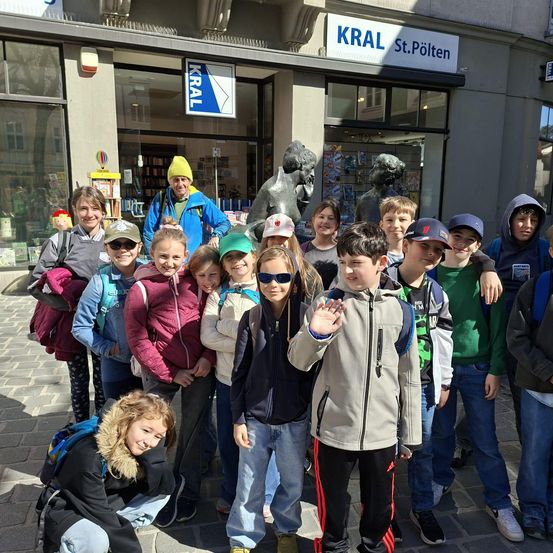 Eine Gruppe von Kindern und Erwachsenen steht vor einem Geschäft mit einer Statue davor. Das Schild des Geschäfts lautet KRAL St. Pölten. Die Kinder lächeln und posieren für ein Foto.
