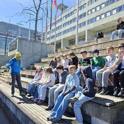 Eine Gruppe von Kindern sitzt auf Stufen vor einem Gebäude. Ein erwachsener Mann steht vor ihnen. Im Hintergrund befindet sich ein Fahnenmast mit Fahnen.