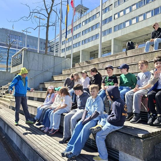 Eine Gruppe von Kindern sitzt auf Stufen vor einem Gebäude. Ein erwachsener Mann steht vor ihnen. Im Hintergrund befindet sich ein Fahnenmast mit Fahnen.