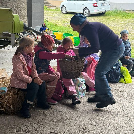 Eine erwachsene Frau gibt einem Kind einen Korb. In der Nähe sitzen Kinder auf Heuballen, und ein weißes Auto ist draußen geparkt.