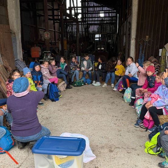 Eine Gruppe von Kindern und Erwachsenen sitzt in einem Kreis in einer Scheune, mit einer Frau vor ihnen, die wahrscheinlich Anweisungen gibt. Verschiedene Taschen und Werkzeuge sind verstreut.