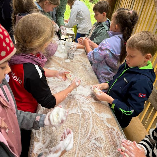 Eine Gruppe von Kindern versammelt sich um einen Tisch, trägt Handschuhe und macht seifige Blasen. Der Tisch ist mit Seifenwasser bedeckt, und die Kinder schauen sich gegenseitig an, während sie Blasen halten.