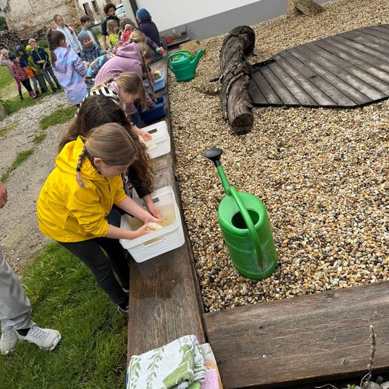 Kinder versammeln sich um eine Holzbank und waschen etwas in einem Behälter mit Wasser. Ein Kind trägt eine gelbe Jacke.