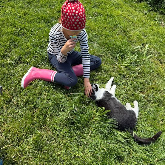 Ein Kind in Stiefeln sitzt auf dem Gras und füttert eine kleine schwarz-weiße Katze. Das Kind trägt eine gepunktete Mütze und hält einen kleinen Gegenstand im Mund.