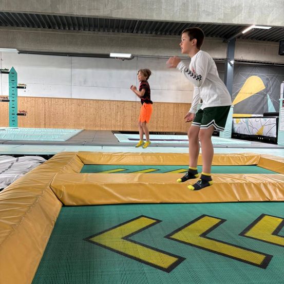 Zwei Jungen springen auf einem Trampolin. Ein Junge trägt ein schwarzes T-Shirt und orange Shorts, der andere trägt ein weißes Langarmshirt und grüne Shorts.