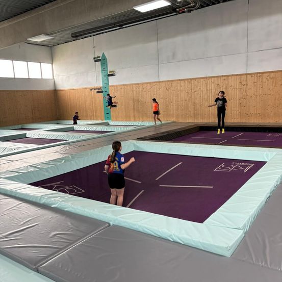 Mehrere Menschen befinden sich in einem großen Indoor-Trampolinpark. Einige springen, während andere zusehen. Die Wände sind aus Holz, und es gibt mehrere Trampolins.