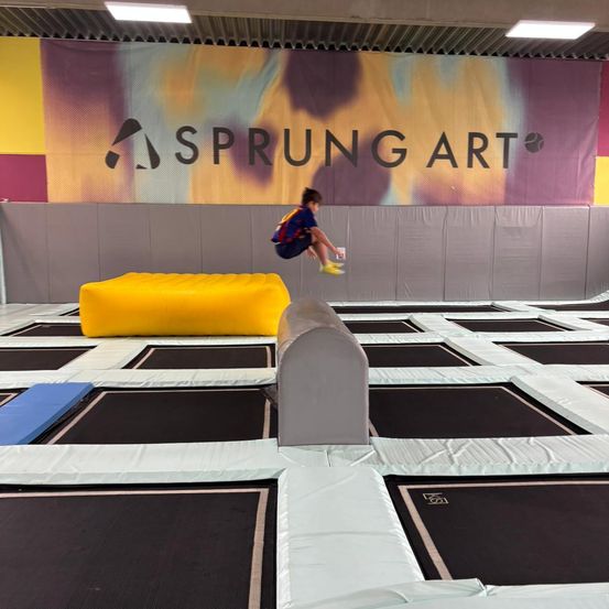 Ein junger Junge springt auf einem Trampolin und überspringt ein gelbes Aufblasbares und ein graues Hindernis. Die Szene spielt sich in einem Sprung Art Indoor-Trampolinpark ab.