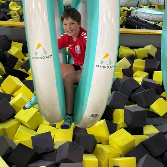 Ein Junge in einem roten Shirt und blauen Socken sitzt in einem aufblasbaren Ring in einem Trampolinpark mit bunten Blöcken.