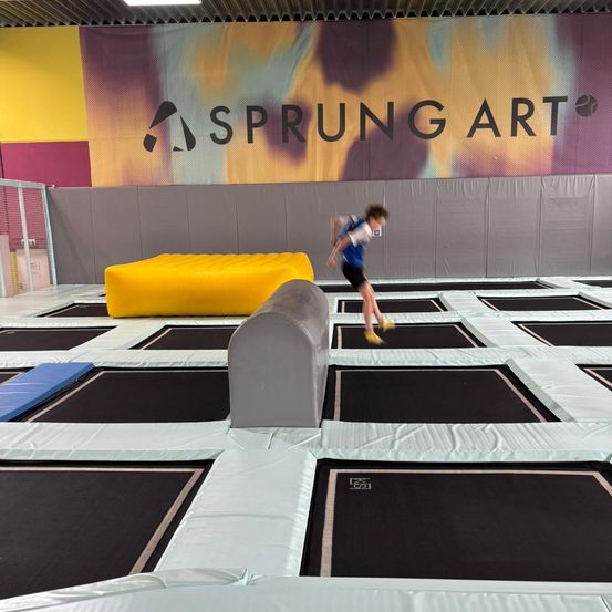 Eine Person läuft auf einem Trampolin mit einem gelben Kissen, trägt ein blaues Shirt, Shorts und gelbe Schuhe. Das Trampolin hat schwarze und graue Abschnitte. Ein Schild mit dem Text 'SPRUNG ART' ist an der Wand.