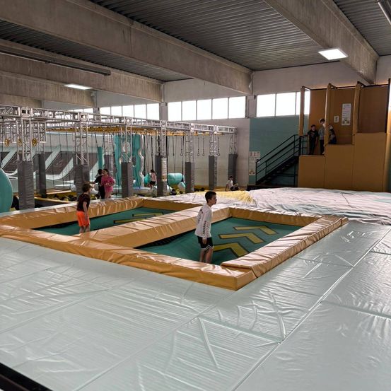 Ein Indoor-Trampolinpark mit verschiedenen Aktivitäten. Ein Junge steht in der Mitte, während andere herumspielen, und es gibt aufblasbare Wände und Klettergeräte.