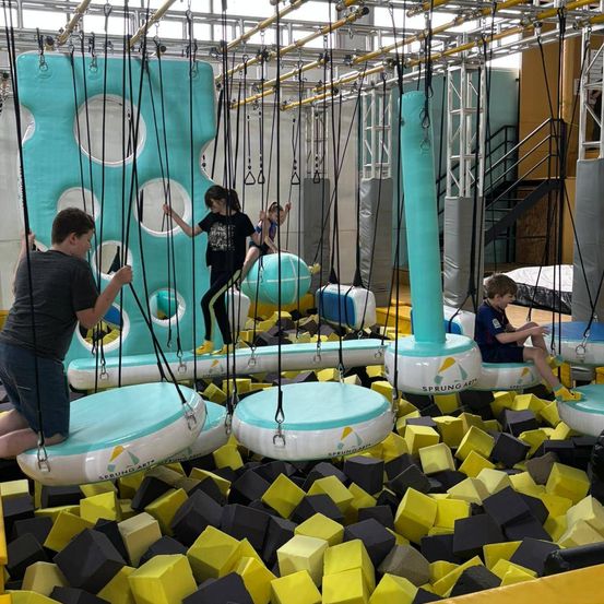 Kinder spielen auf Schaukelsets in einem Trampolinpark. Einige hängen an Hängeschaukeln, andere sitzen auf hängenden Plattformen. Bunte Schaumstoffblöcke bedecken den Boden.
