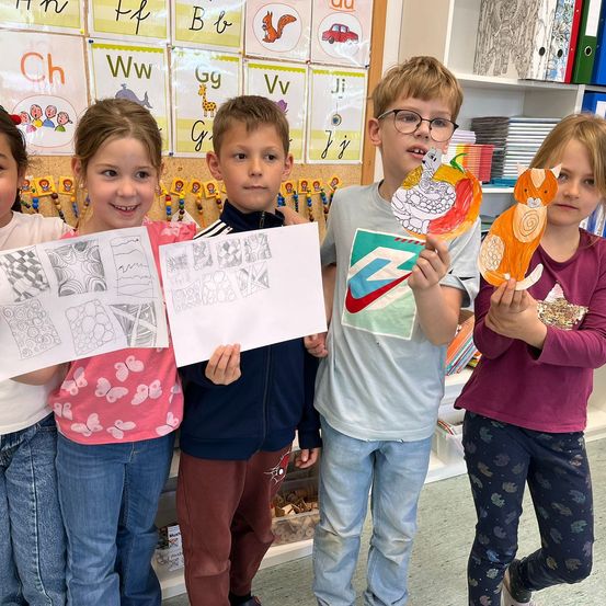 Eine Gruppe von Kindern in einem Klassenzimmer posiert für ein Foto. Sie halten Zeichnungen und Bastelarbeiten in den Händen. Die Wände sind mit Alphabettafeln und Buchstabenkarten dekoriert.